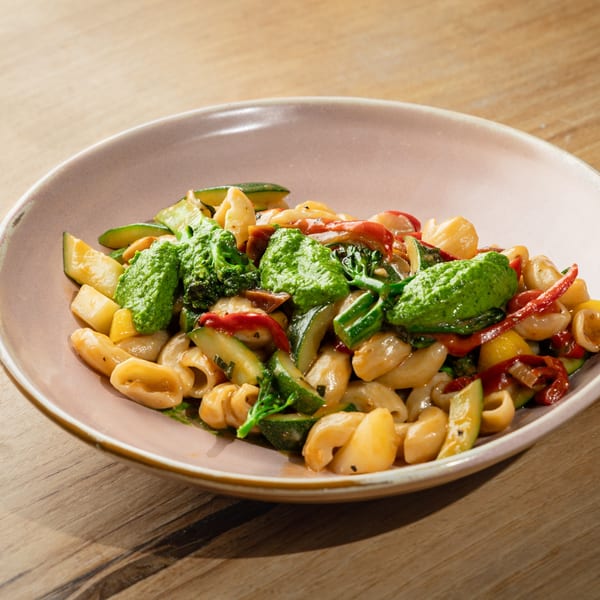 Orecchiette pasta with zucchini, red peppers, broccoli, and a dollop of green pesto in a pink bowl.