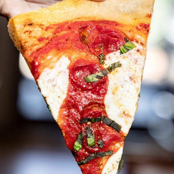 A hand holds up a slice of Earl & Isabella's Taverna's Italian pizza with tomato sauce, mozzarella, and basil.
