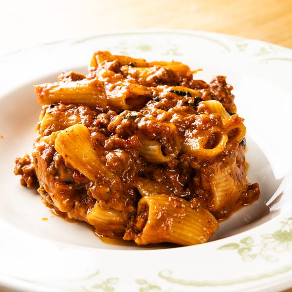 Close-up of rigatoni pasta with a rich, chunky red meat sauce in a white bowl.