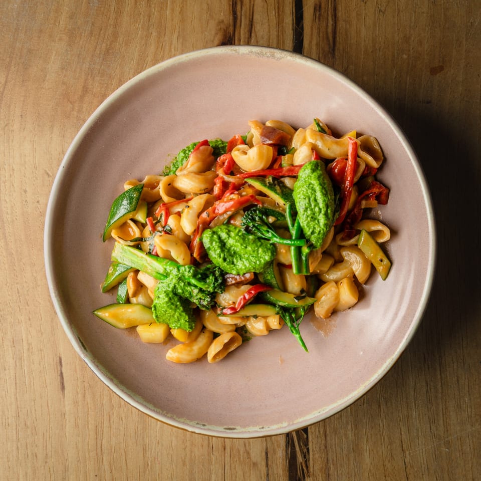 Italian pasta dish with pesto, red peppers, zucchini, and broccoli on a pink plate.