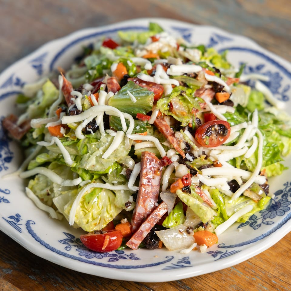 Italian salad with lettuce, salami, cheese, tomatoes, olives, and carrots on a patterned plate.