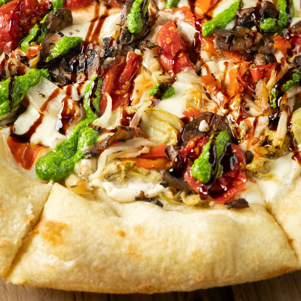 Close-up of an Italian pizza with pesto, balsamic glaze, mushrooms, tomatoes, and melted cheese.