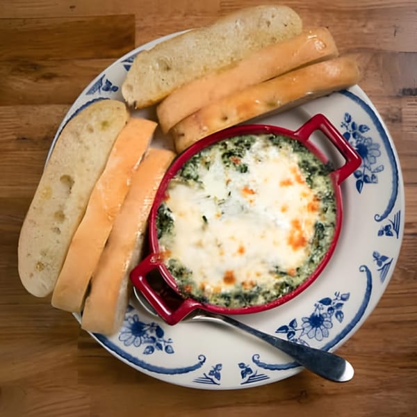 Baked spinach dip in a red dish with toasted breadsticks and sliced bread on a decorative plate.